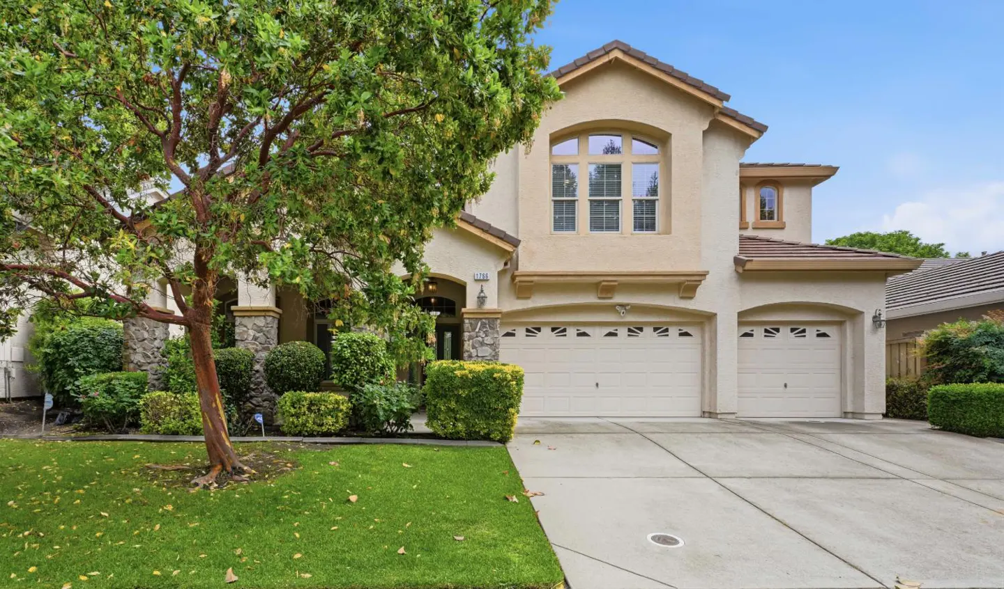 Two-story beige house with a two-car garage, green lawn, and a large tree in the front yard under a blue sky.