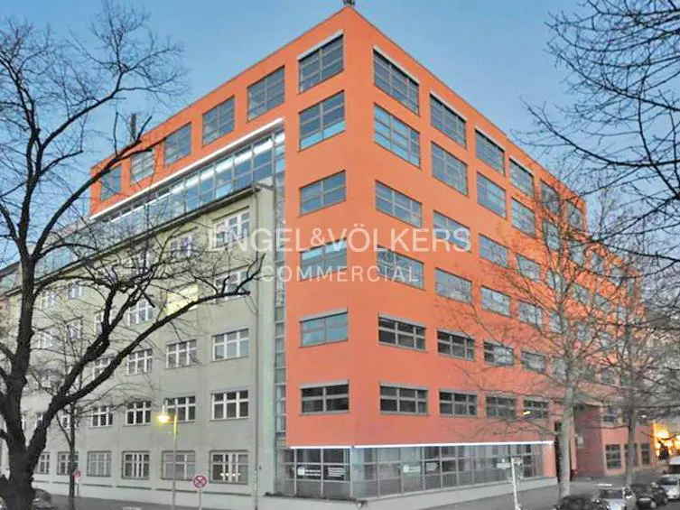 A modern corner office building with a bright orange facade and a grey lower section, framed by bare trees against a sky.