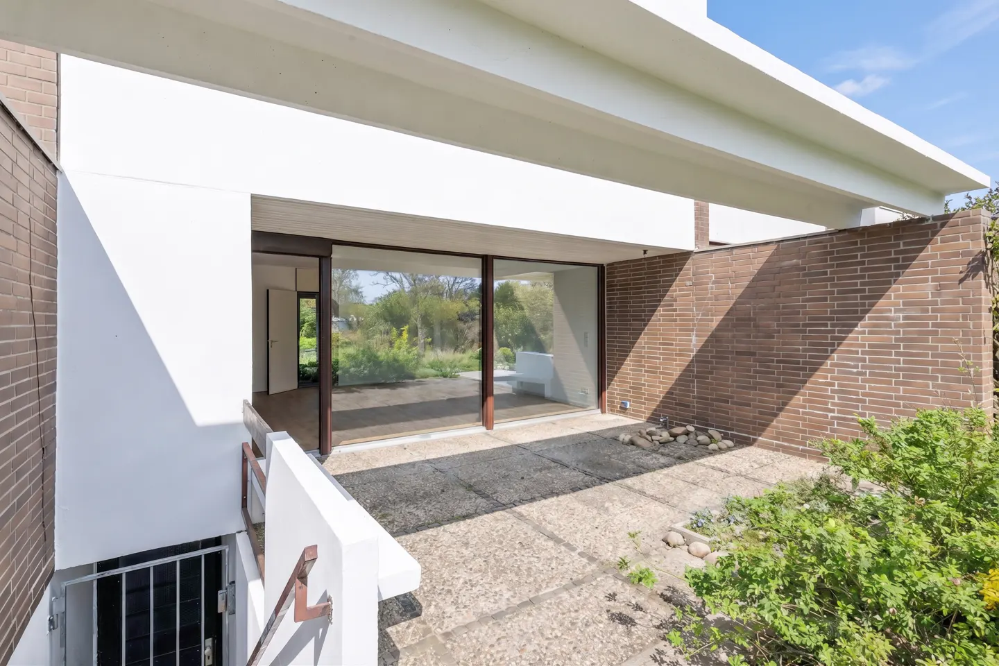 Exterior view of a modern home with brick and white walls, large glass sliding doors, and a concrete patio. A gated entrance leads to a lower level.