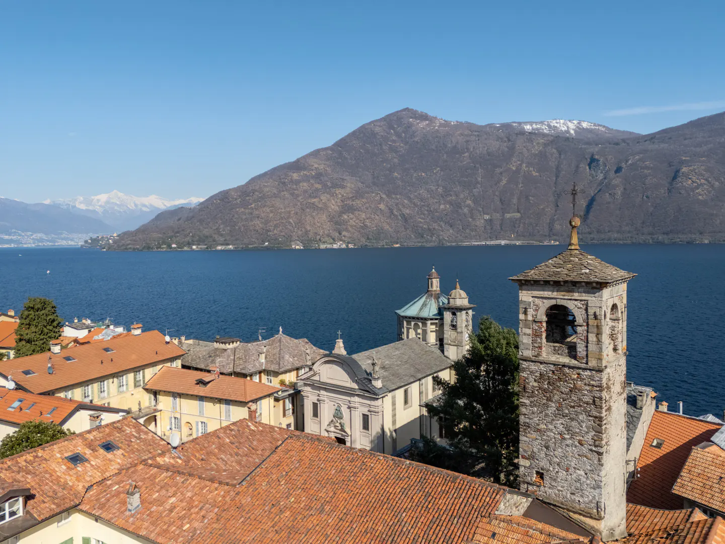 Scenic view of Lake Maggiore, Italy, with terracotta rooftops, a stone church tower, and mountains in the background under a clear blue sky.