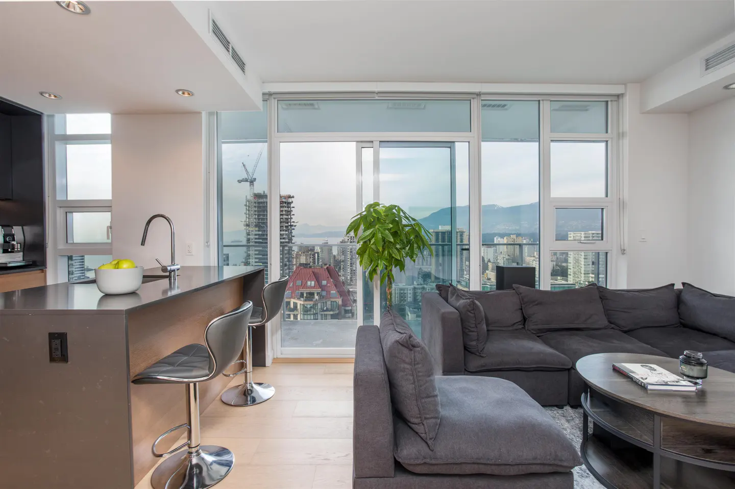 Modern condo interior with a gray sectional sofa, kitchen island with stools, and a large window overlooking a city skyline.