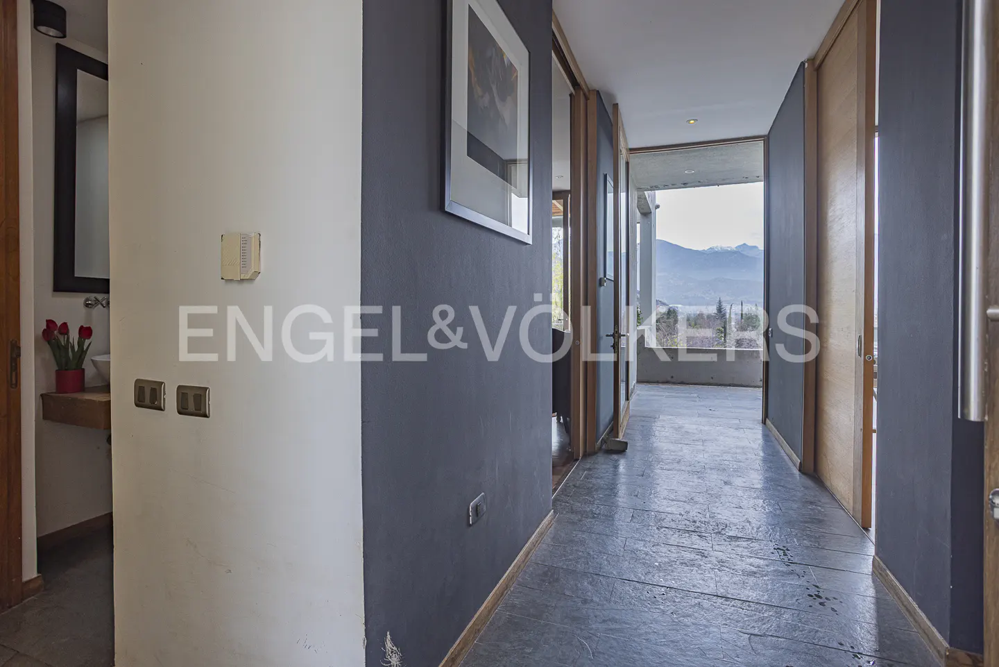 Modern hallway with dark gray walls, stone flooring, and a view of distant mountains through a glass-walled balcony.