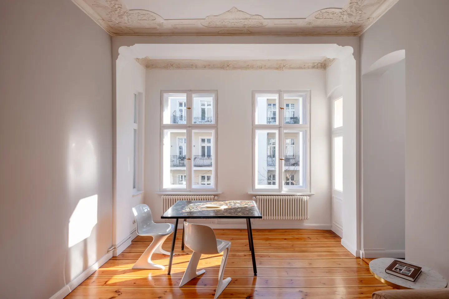Bright, empty room with wood floors, white walls, and ornate ceiling. A table and two chairs sit in front of two large windows.