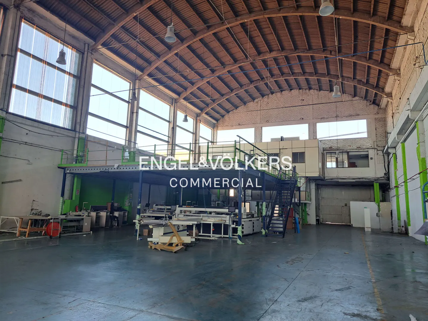 A large, empty warehouse with a high, arched wooden ceiling and concrete floors. Natural light streams in through tall windows. Machinery is visible in the foreground.