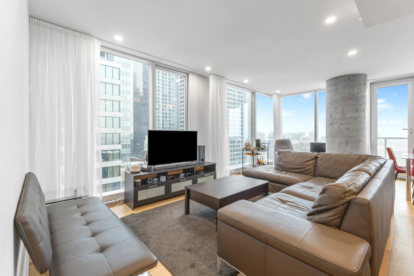 Modern living room with a grey sectional sofa, coffee table, TV console, and floor-to-ceiling windows overlooking a city.