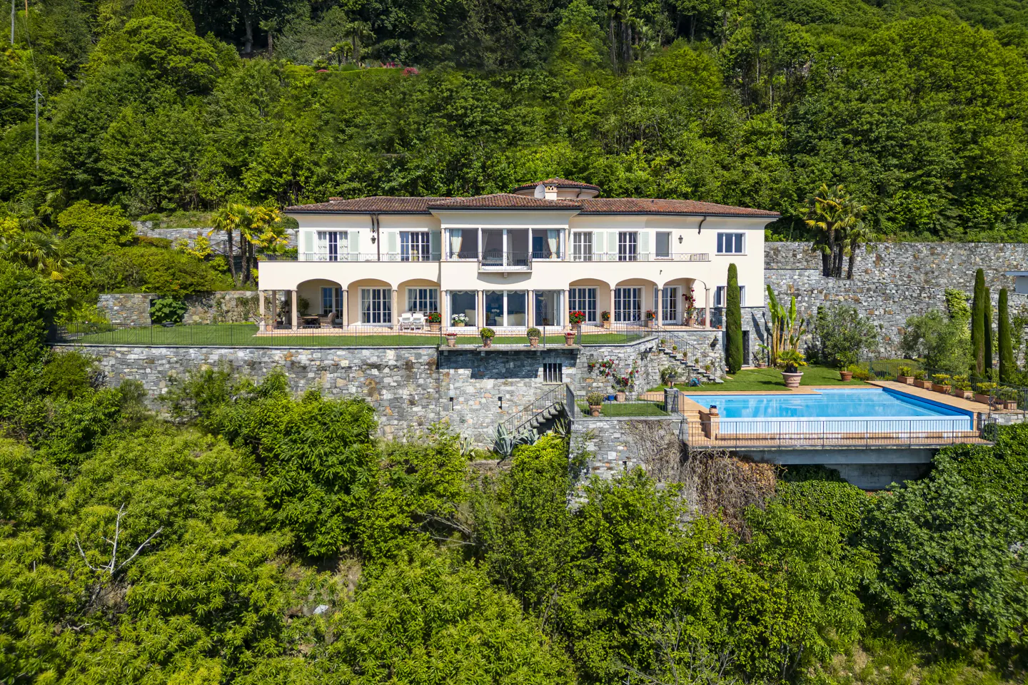 A cream-colored villa with arched terraces sits on a stone hillside surrounded by lush trees, featuring a private pool.
