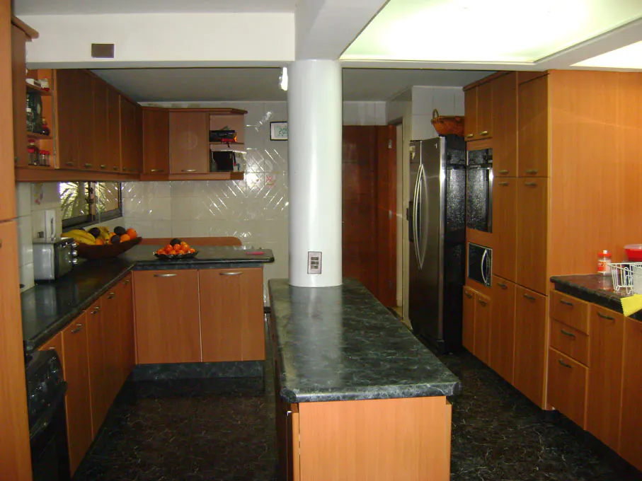 A kitchen with wood cabinets, black countertops, and a white pillar in the center. A stainless steel refrigerator is visible.
