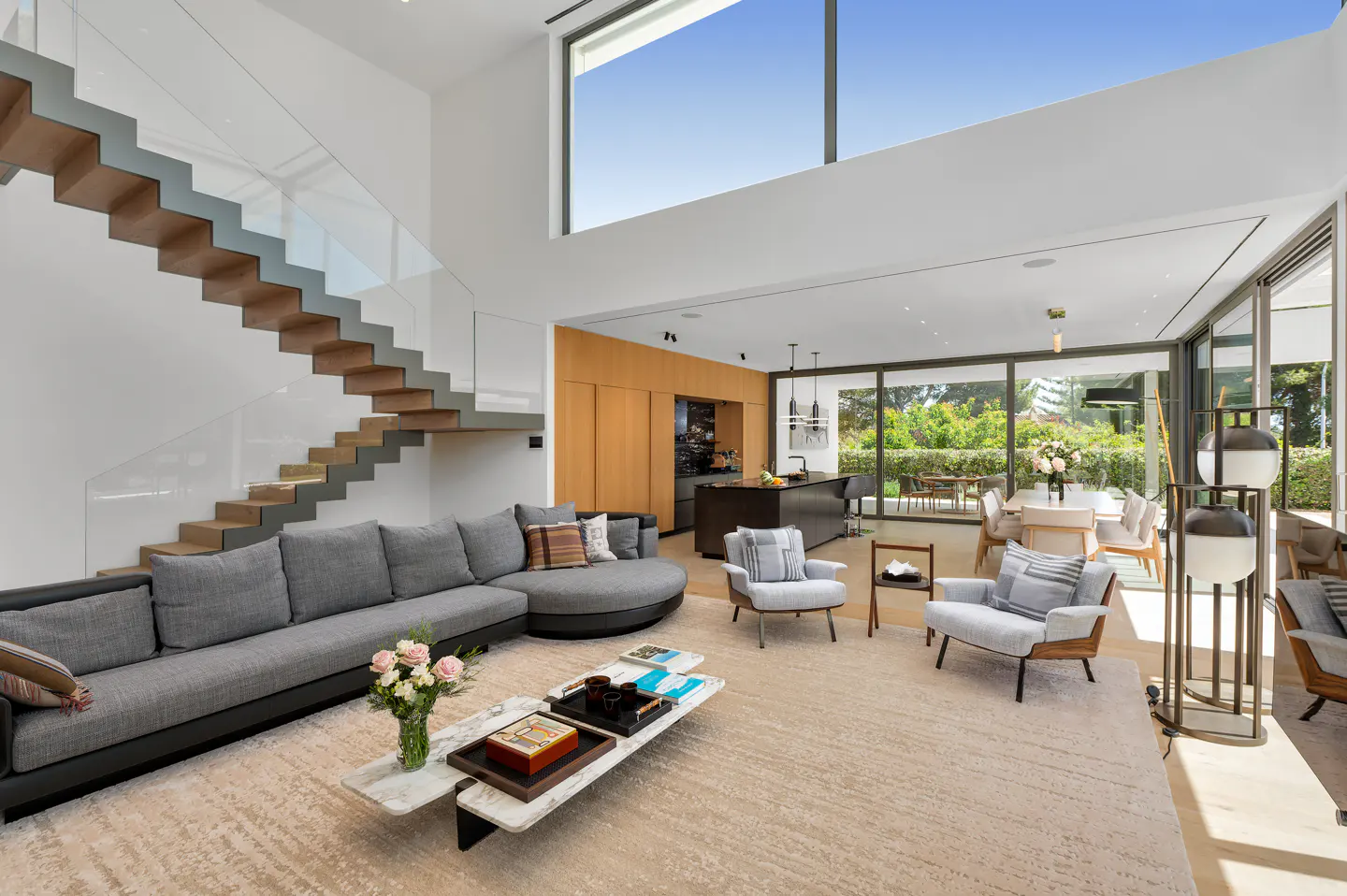 Open-concept living room with gray sofa, floating stairs, and floor-to-ceiling windows. Kitchen and dining area visible in background.