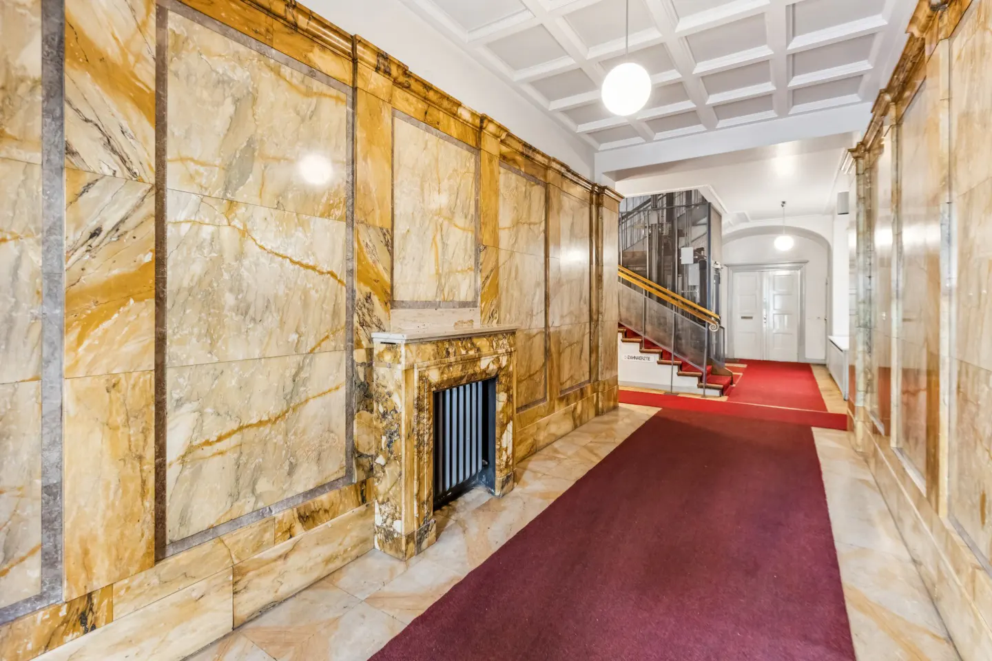 Hallway with marble walls, red carpet, and coffered ceiling. A staircase and elevator are visible in the background.