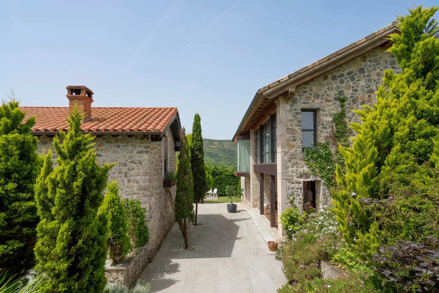 Exterior view of two stone buildings with red tile roofs, separated by a stone walkway and green trees.