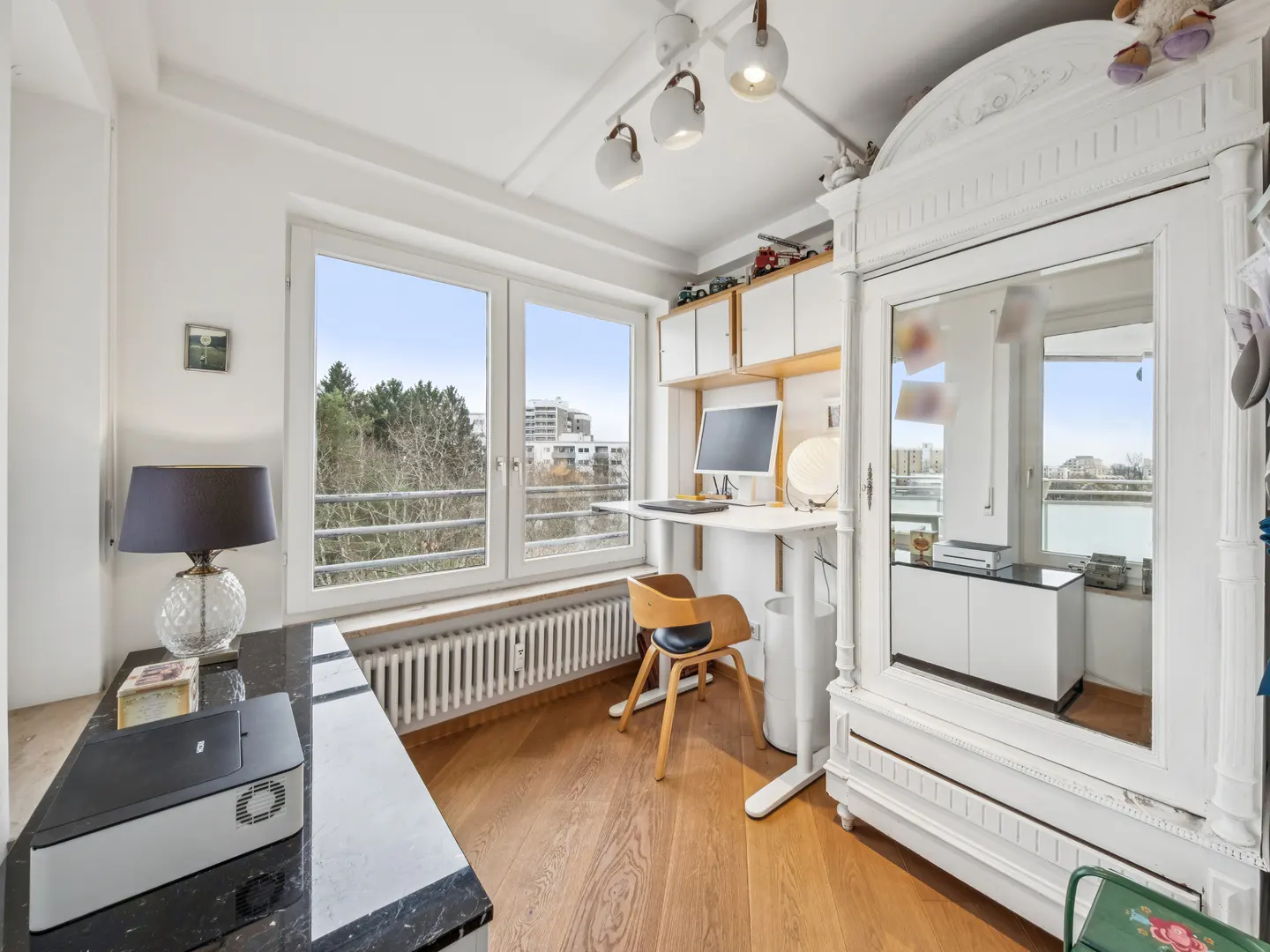 Bright room with wood floors, a white standing desk, and a large window with a city view. A black marble table and white wardrobe are also visible.