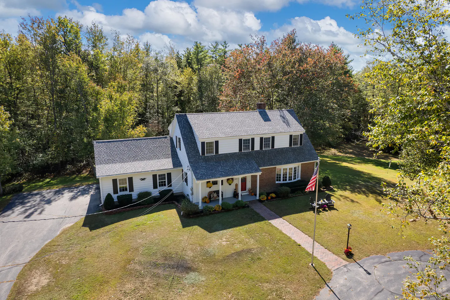 Aerial view of a two-story white house with black shutters, a gray roof, and a brick facade, surrounded by trees.