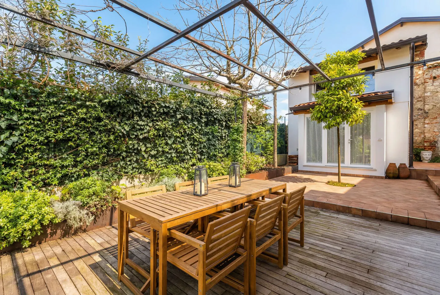 Outdoor dining area with a wooden table and chairs under a metal pergola, surrounded by lush greenery and a white house.