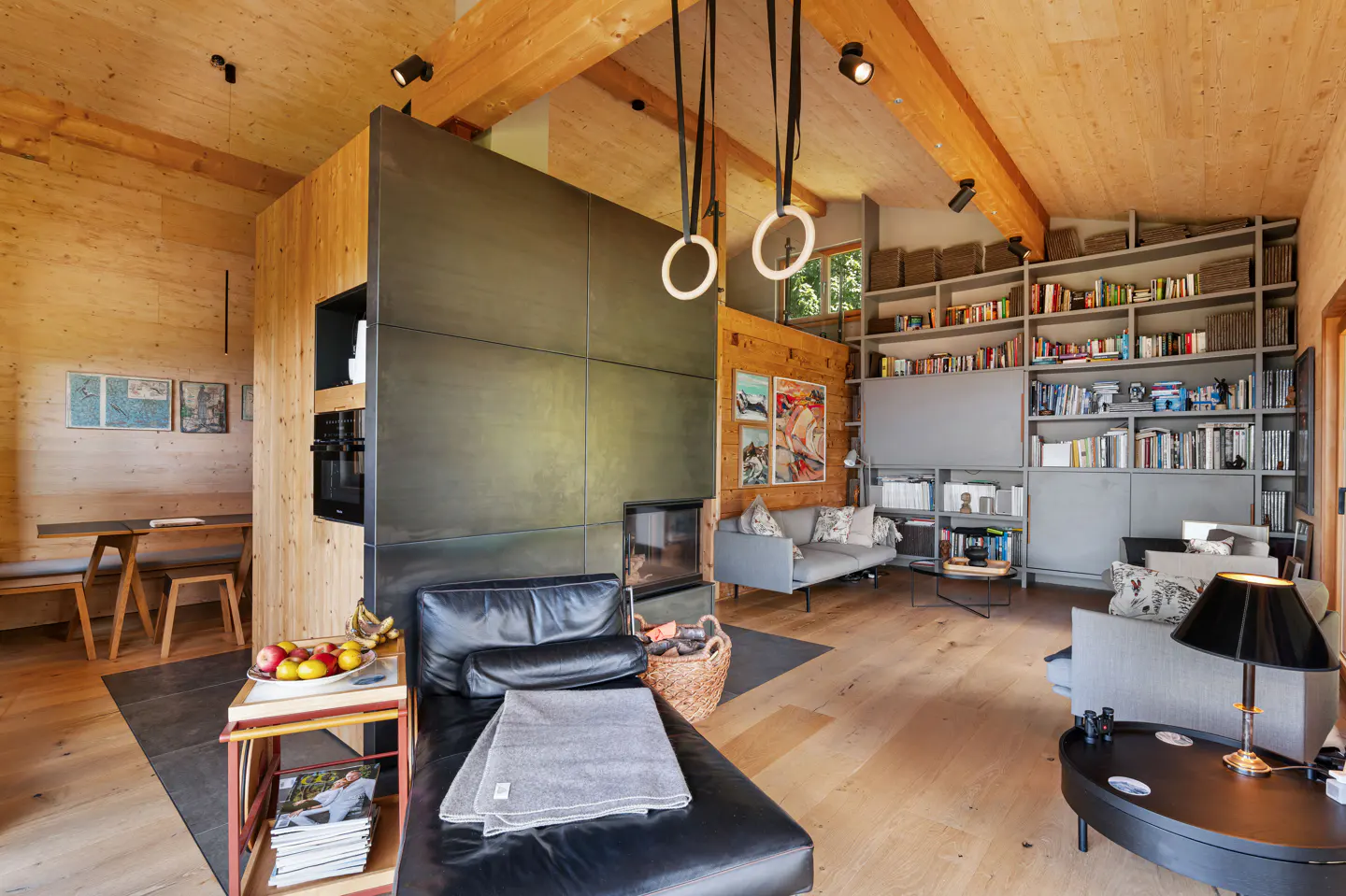 A modern living room with wood walls, beams, and a black leather chaise lounge. A bookshelf is filled with books and baskets.