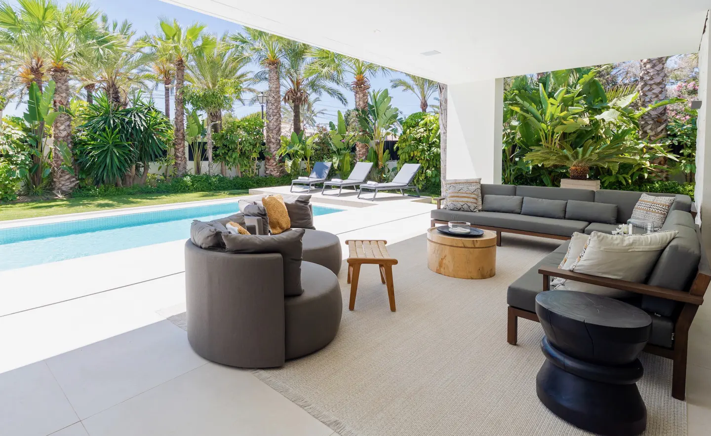 Outdoor patio with gray sofas, wood tables, and a pool. Palm trees and greenery in the background.
