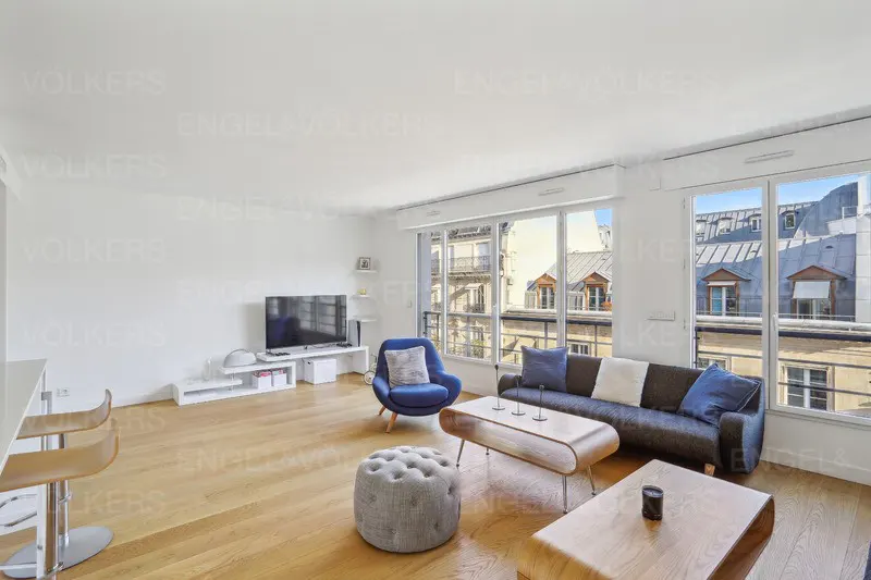 Bright living room with wood floors, white walls, and large windows showing city views. A blue chair, gray sofa, and modern tables furnish the space.