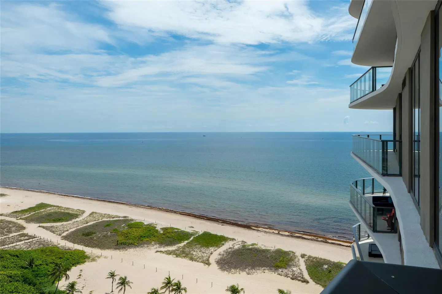 View from a high-rise balcony overlooking a sandy beach, blue ocean, and partly cloudy sky. Palm trees are visible on the beach.