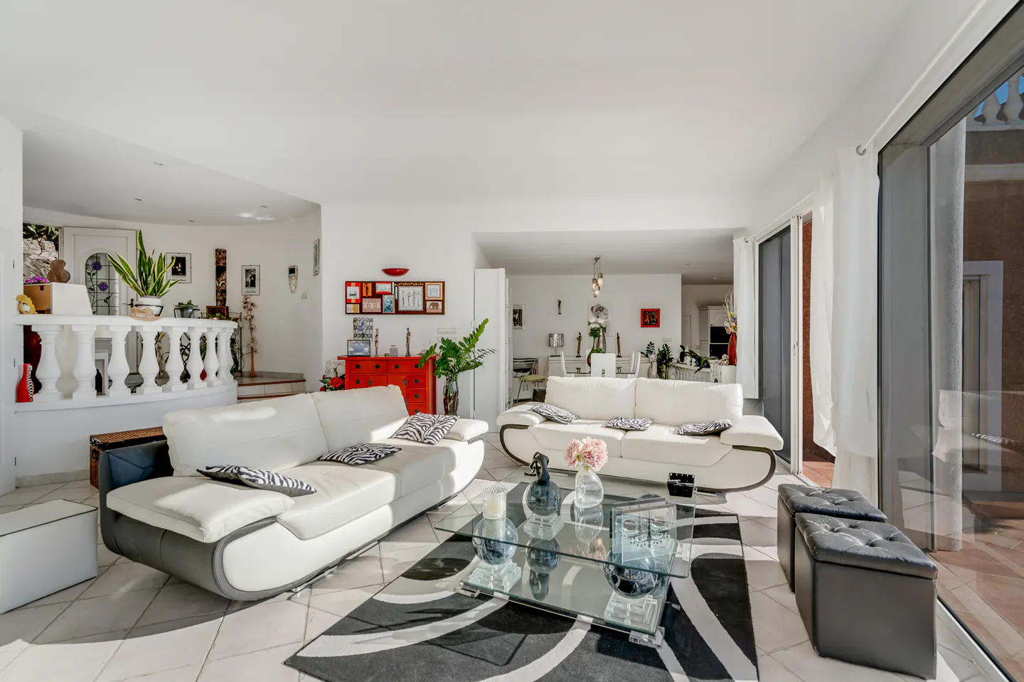 Bright living room with white sofas, glass table, and black/white rug. Balcony visible through sliding glass doors.
