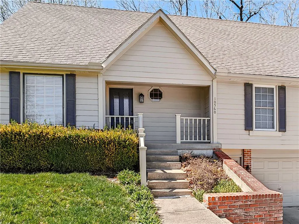 Beige house with gray shutters and a gray roof. A walkway leads to steps up to a covered porch with white railings.