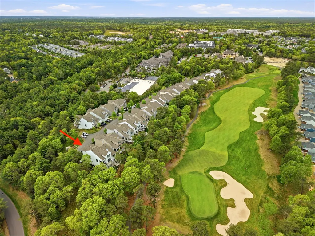 Aerial view of townhouses next to a golf course, surrounded by green trees. An arrow points to one of the townhouses.