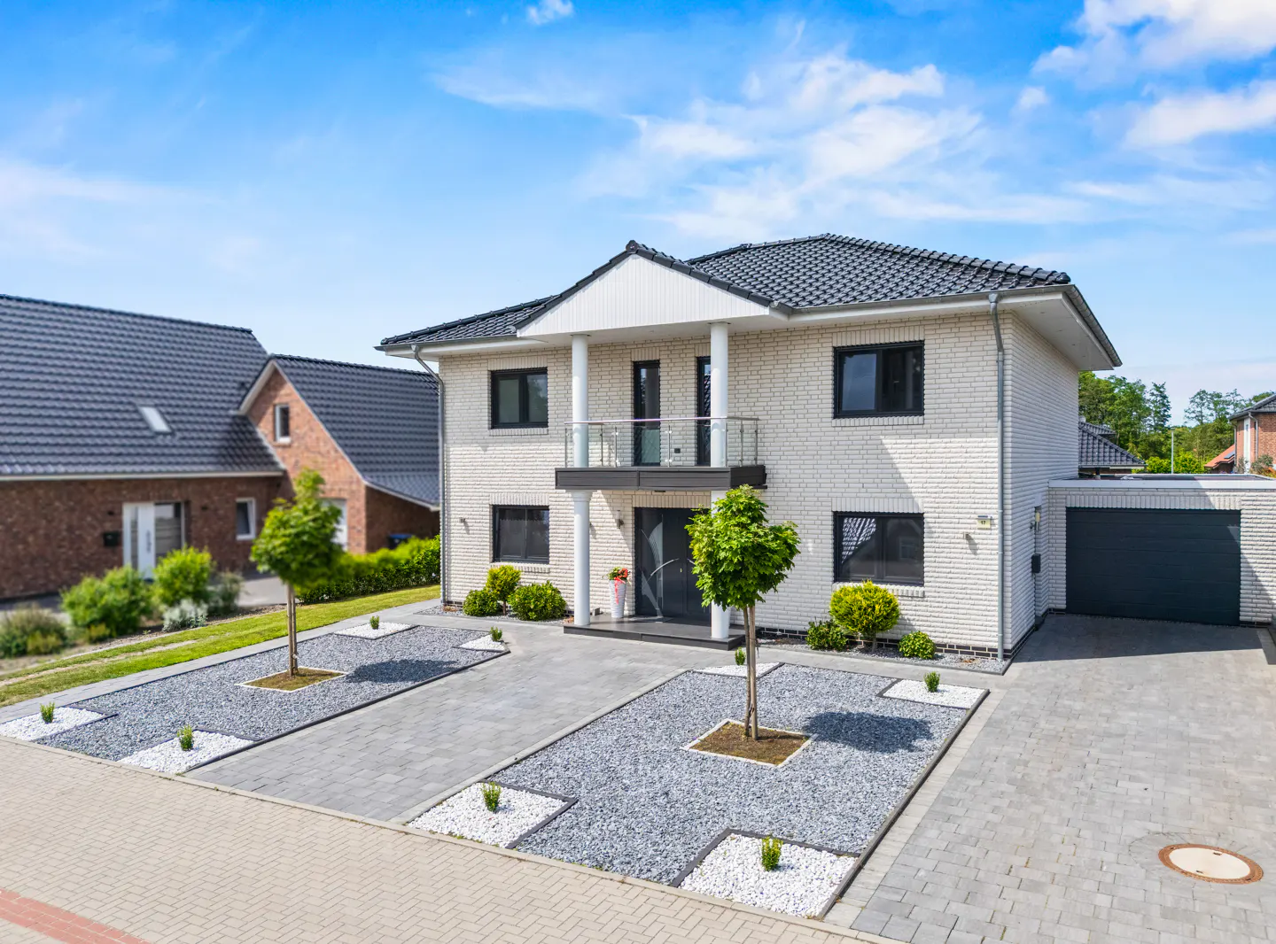 Modern two-story white brick house with a dark roof, balcony, and a paved front yard with small trees and gravel beds.