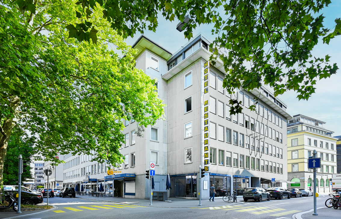 A multi-story white hotel building with a vertical "HOTEL RESTAURANTS" sign, framed by green trees on a city street corner. [AI-generated text]