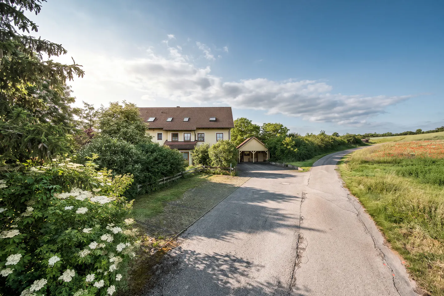 A two-story house with a brown roof and a carport sits beside a country road, surrounded by lush greenery and a field.