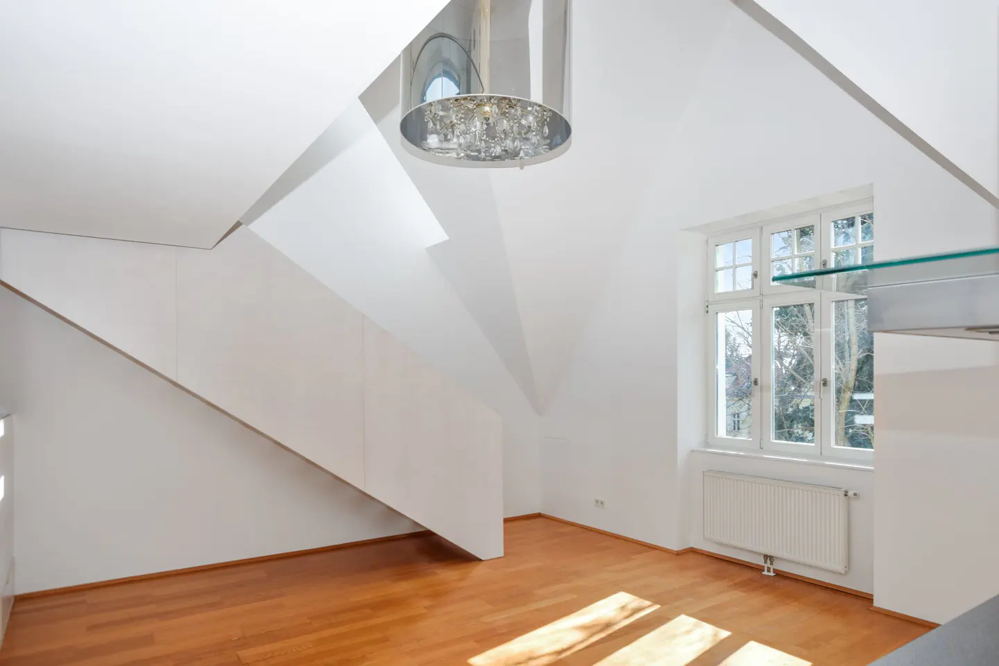 Bright, modern room with hardwood floors, white walls, and a staircase. A chandelier hangs from the ceiling, and a window lets in natural light.
