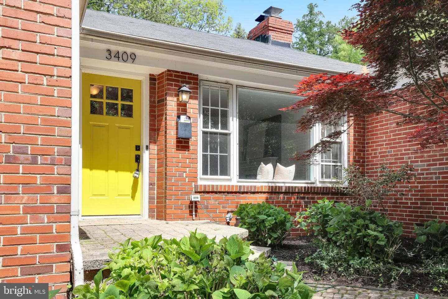 Red brick house with a bright yellow front door, a large window, and green landscaping. Address number 3409 is visible above the door.