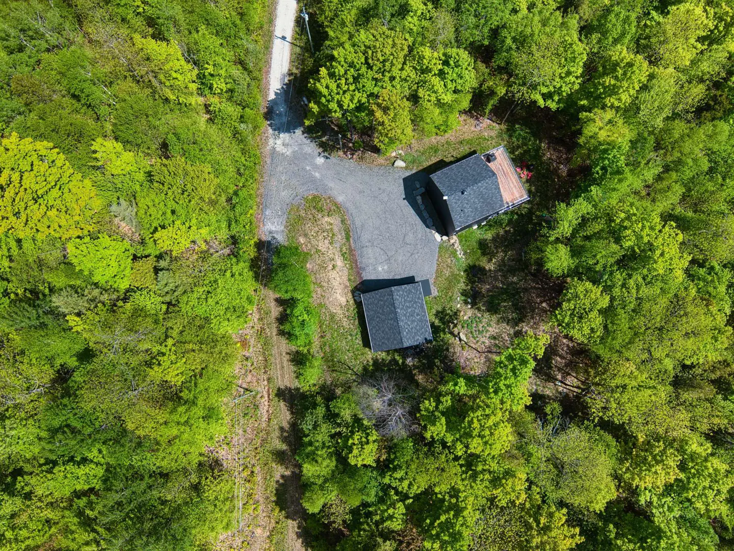 Aerial view of two modern, dark-colored houses with gray roofs nestled in a lush green forest, connected by a gravel driveway to a road.