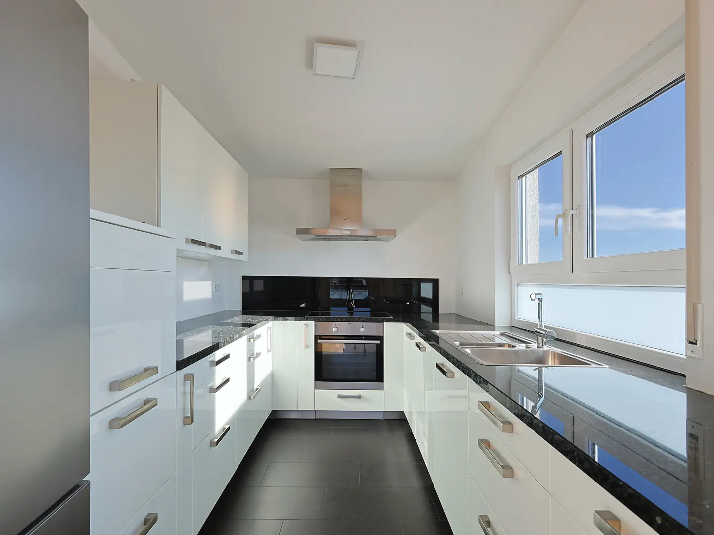 Bright kitchen with white cabinets, black countertops and backsplash, stainless steel appliances, and a window with a blue sky view.