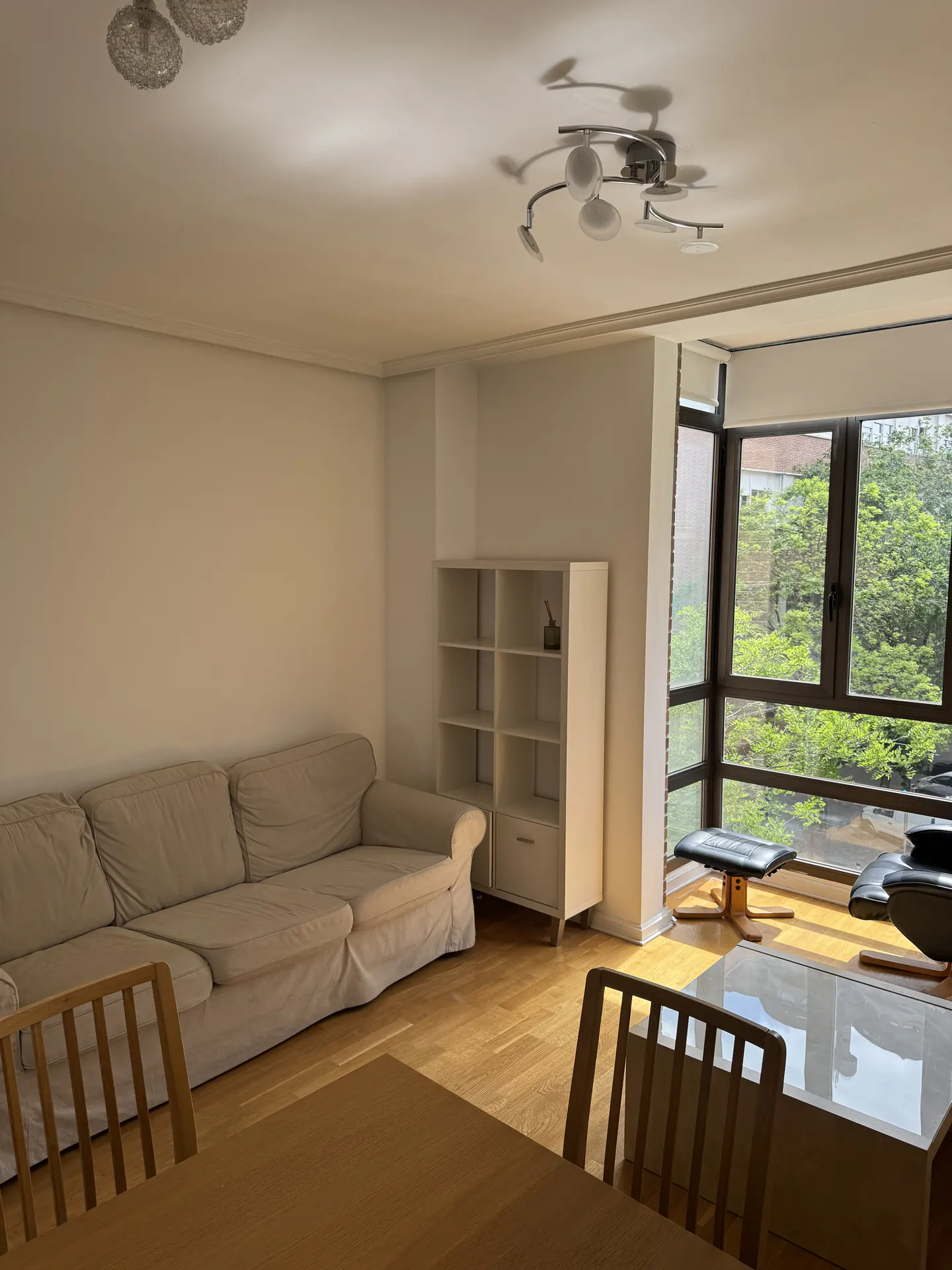 Bright living room with a beige sofa, wooden table and chairs, white bookshelf, and large windows overlooking green trees.
