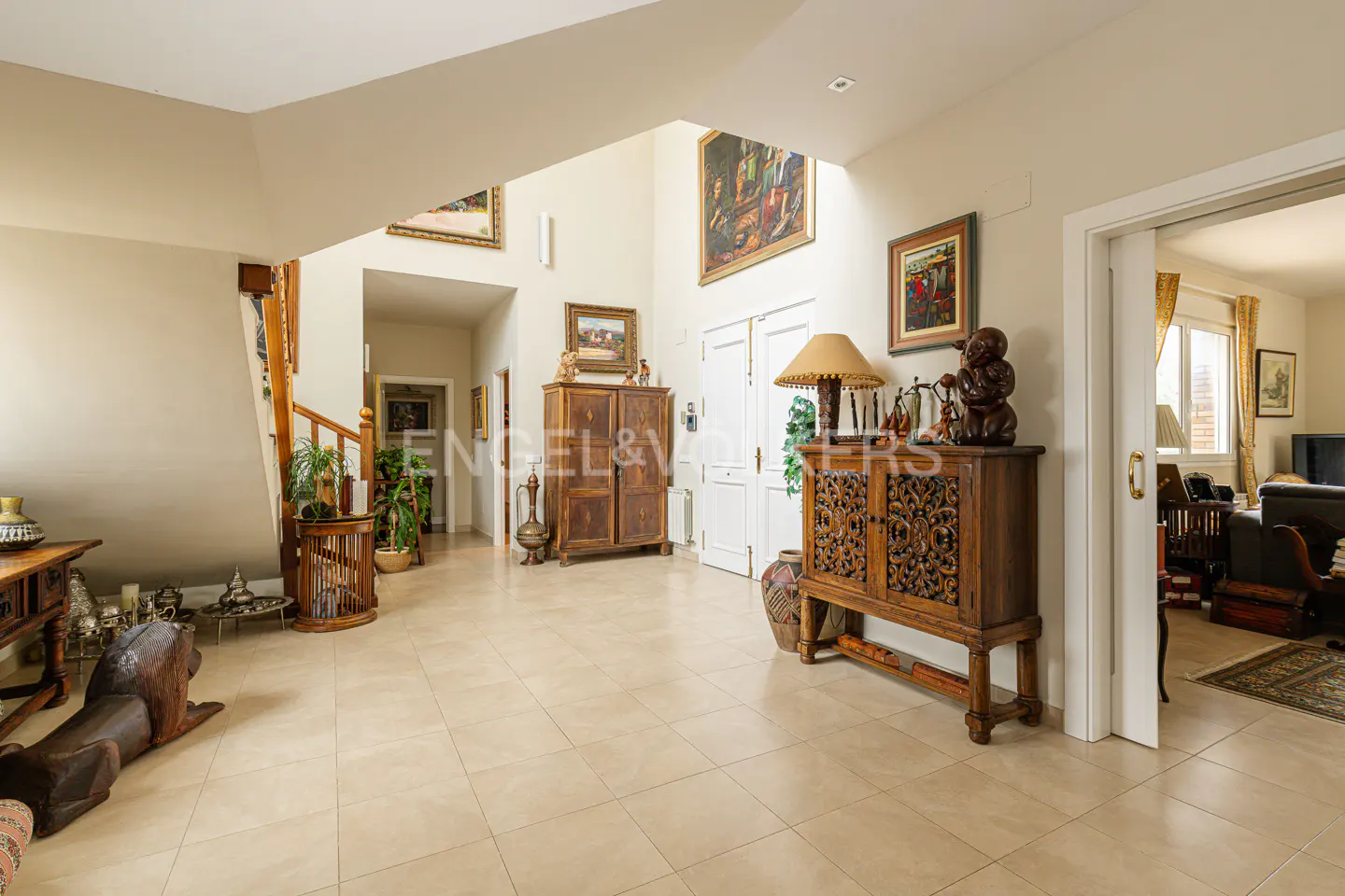 A bright foyer with beige tile floors, wooden furniture, and artwork on the walls. A staircase is visible on the left.