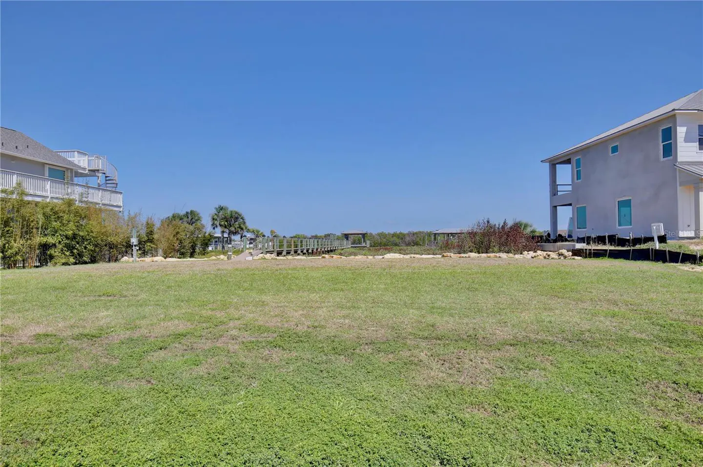 Vacant lot with green grass, between two houses, under a clear blue sky. A wooden boardwalk is in the background.