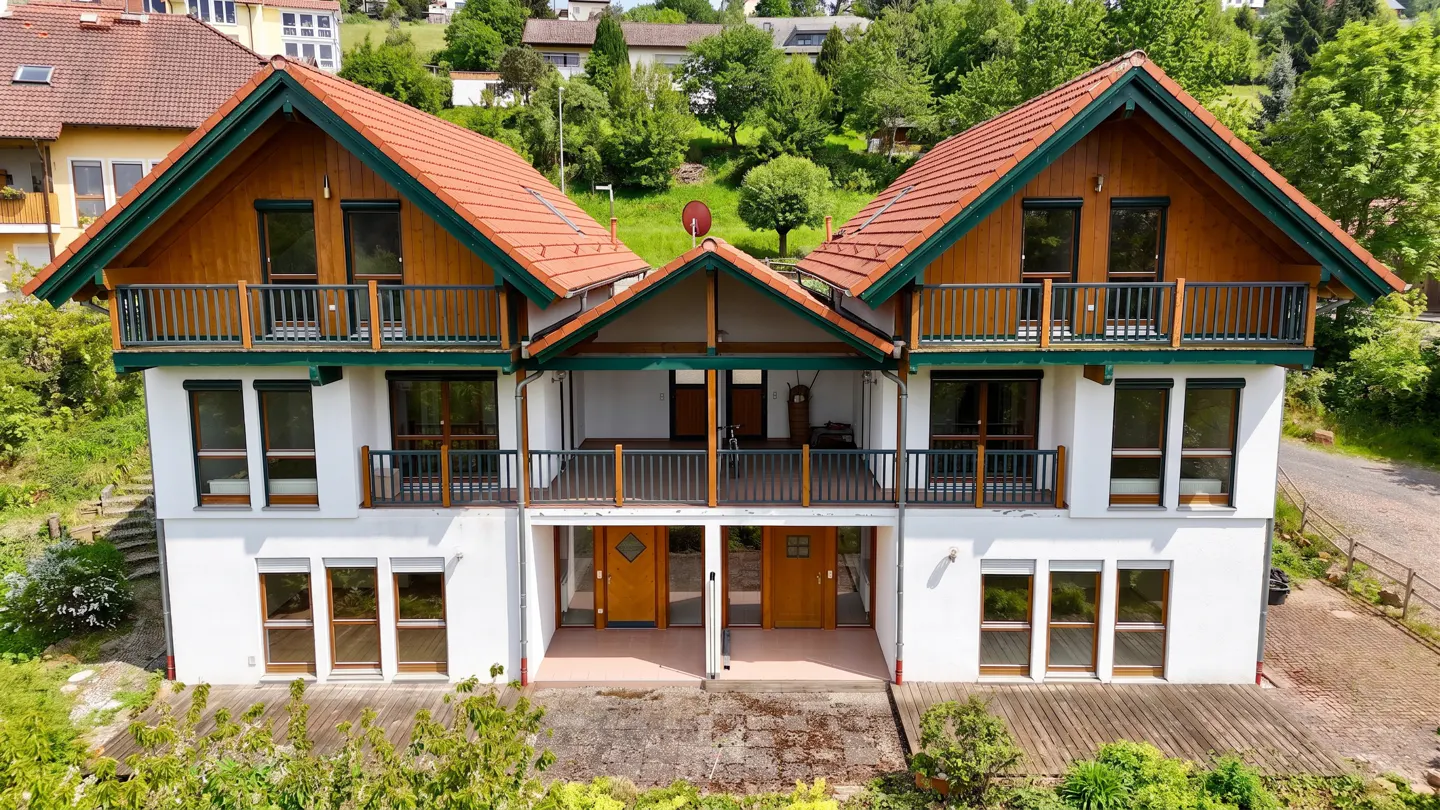 Two-story duplex with orange tile roof, wood siding, and white stucco. Balconies on the second floor with green trim. Lush green hillside in background.