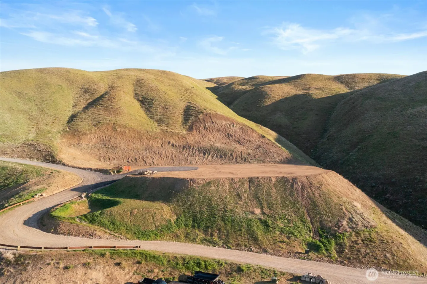 Rolling green hills under a blue sky. A dirt road winds up to a cleared, flat area, possibly a building site.