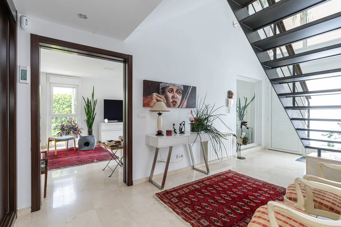 Bright foyer with marble floors, a red patterned rug, and a white console table with a lamp and art. A black staircase is on the right.