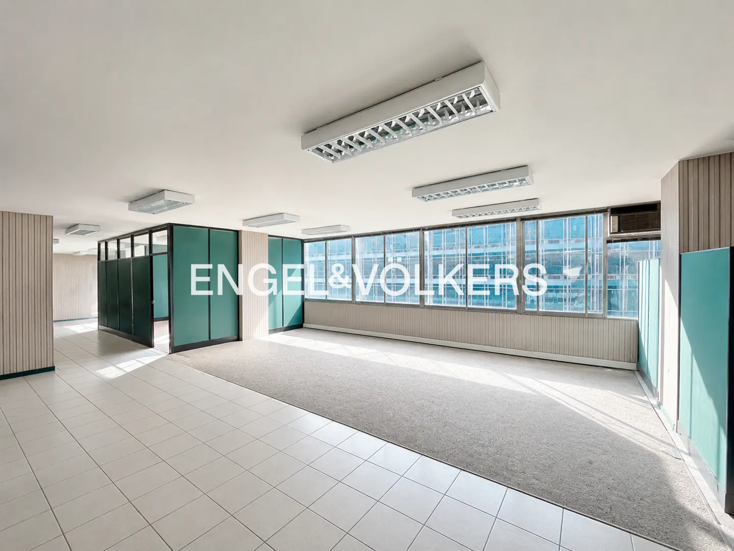 Empty office space with white tiled floors, a large carpeted area, teal partitions, and windows overlooking a city street.