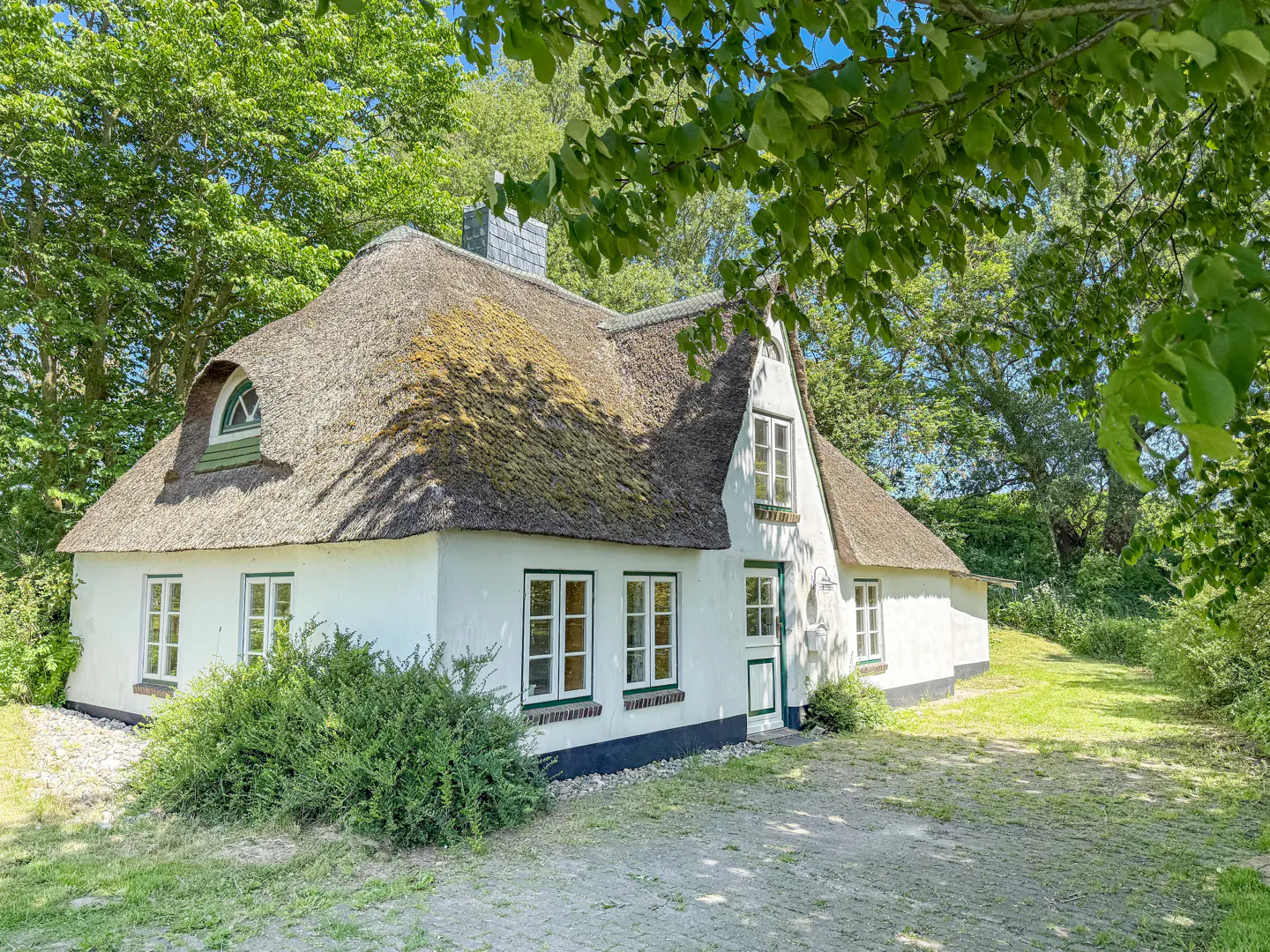 Charming white cottage with a thatched roof, green trim, and lush trees surrounding it.