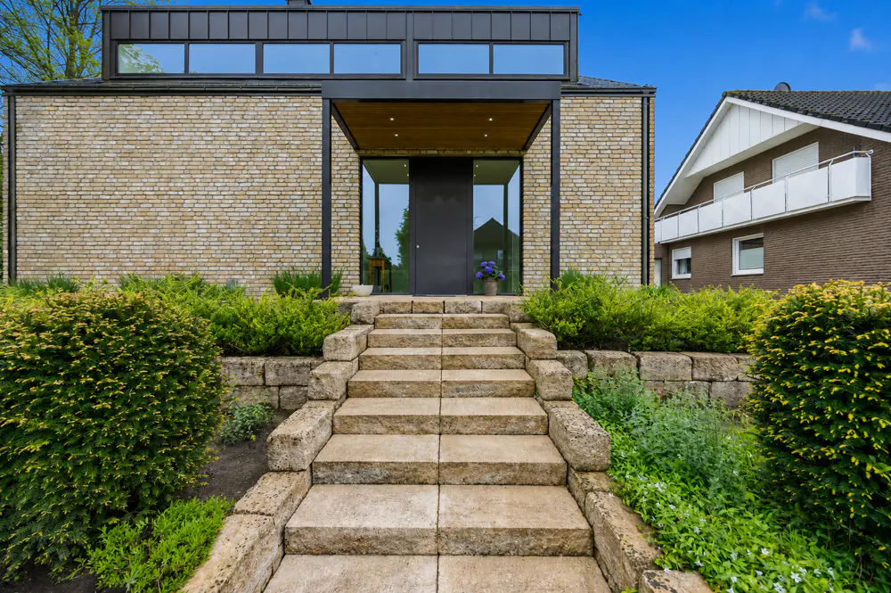 Modern home exterior with stone steps leading to a black front door. The house has a brick facade and large windows. Green bushes line the walkway.