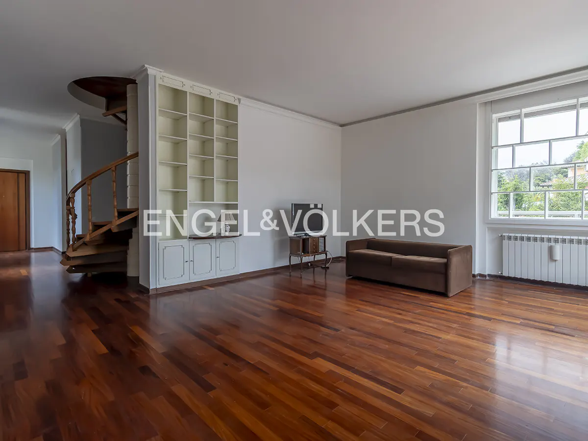 Bright, empty living room with hardwood floors, spiral staircase, built-in shelves, brown sofa, and large window.