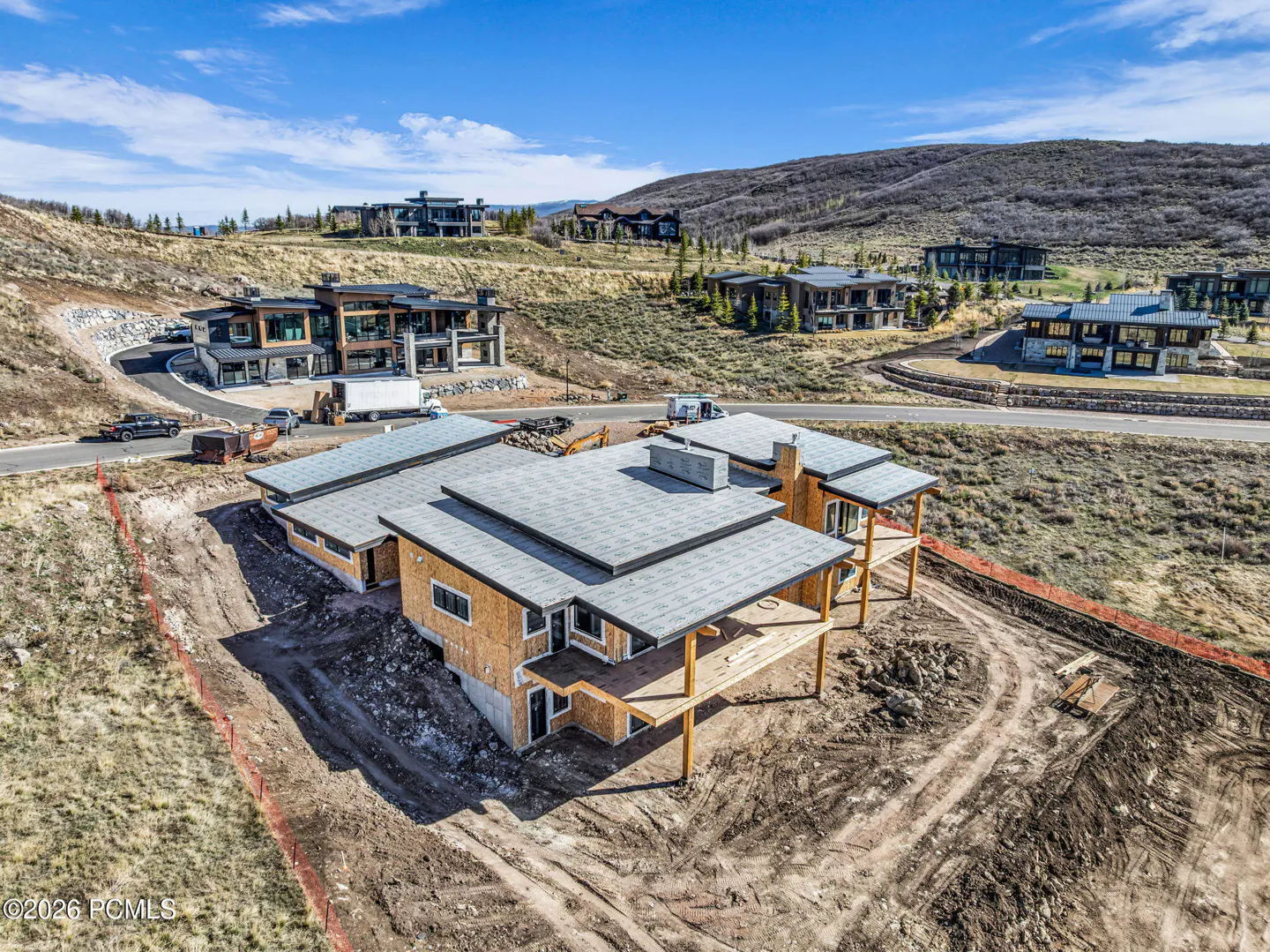 Unfinished home with gray roof and wood frame on a dirt lot. Other modern homes are on a hillside in the background under a blue sky.