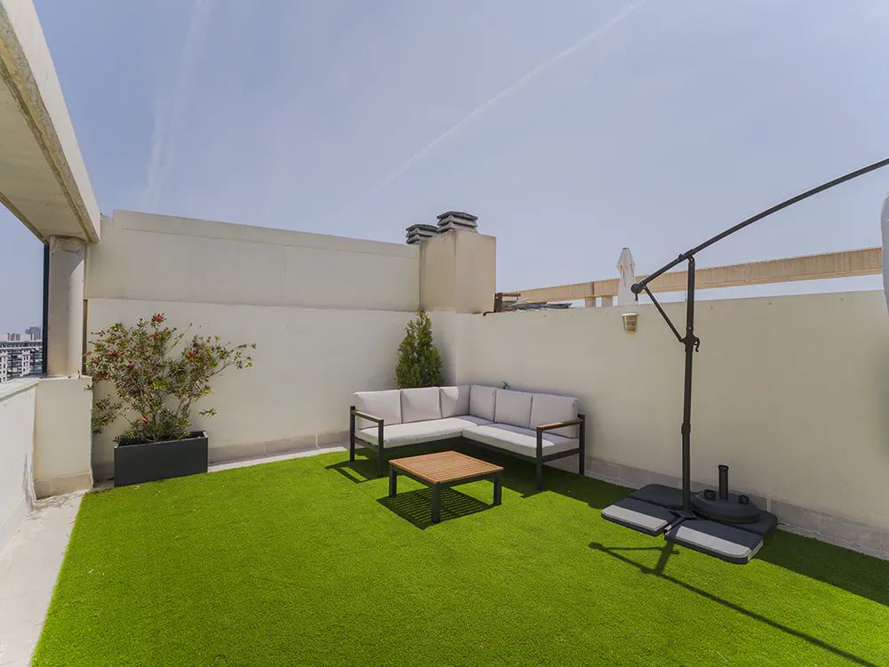 Rooftop patio with green turf, white walls, and blue sky. Features a gray sectional sofa, wood table, potted plants, and umbrella.