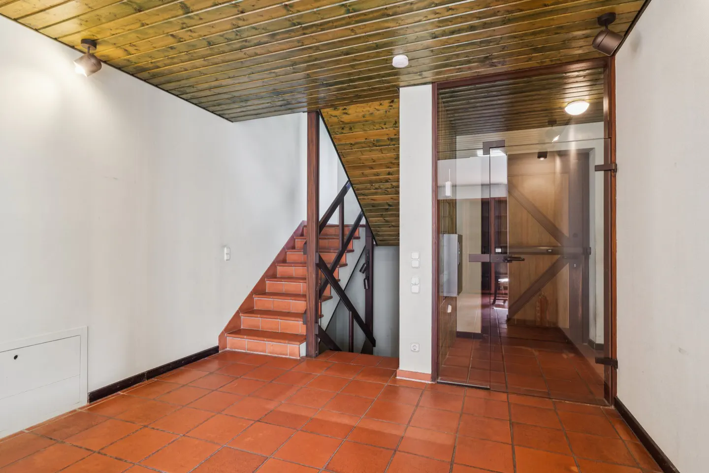 Interior view of a home's entryway with terracotta tile floor, wooden ceiling, stairs, and glass door.