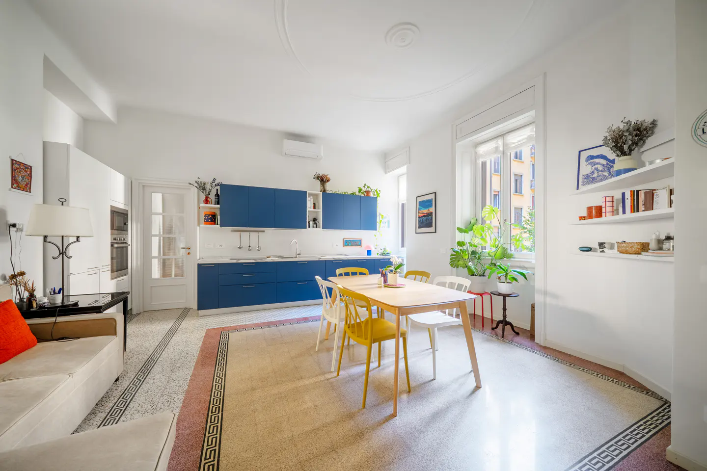 Bright, modern kitchen with blue cabinets, a light wood table with yellow and white chairs, and a large window with plants.
