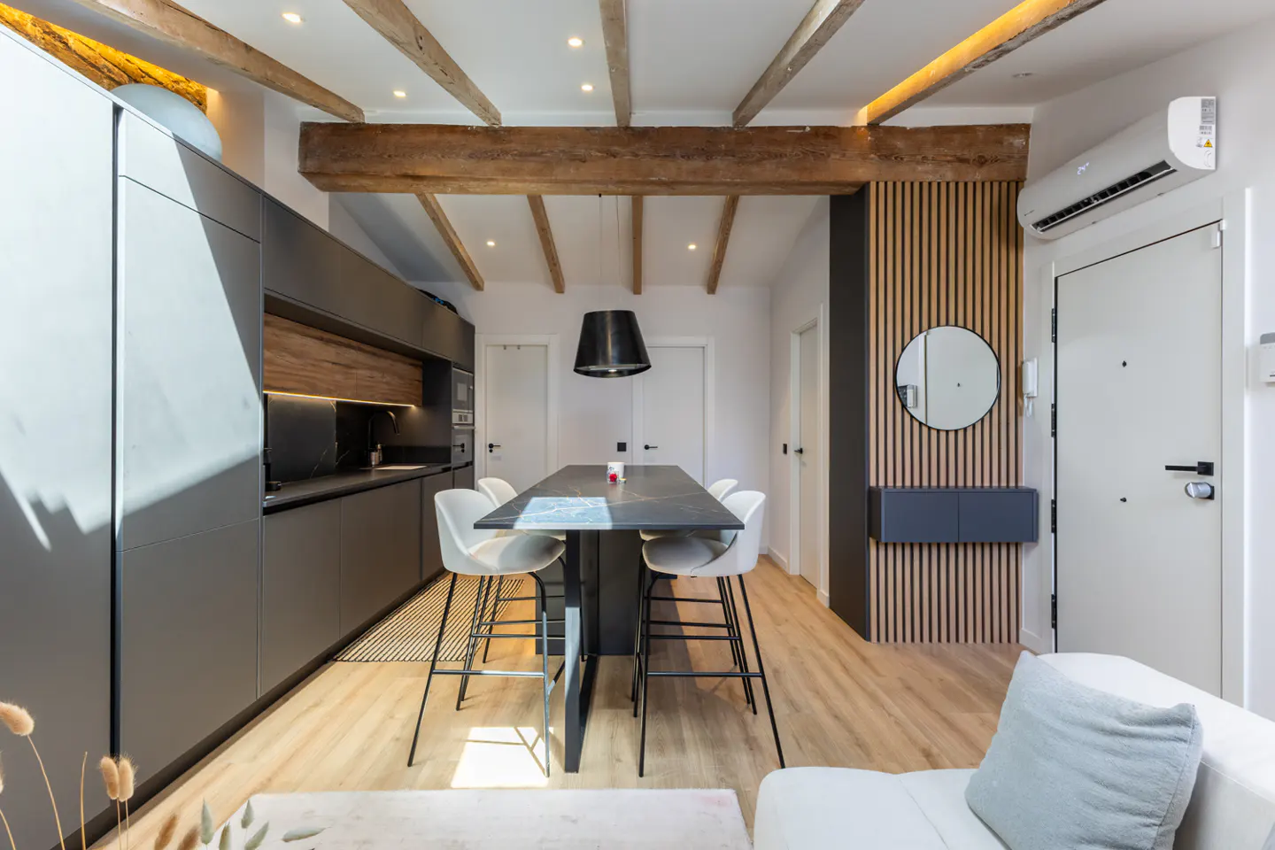 Modern kitchen and dining area with gray cabinets, wood beams, and a black marble table with white chairs. A round mirror hangs on a wood slat wall.