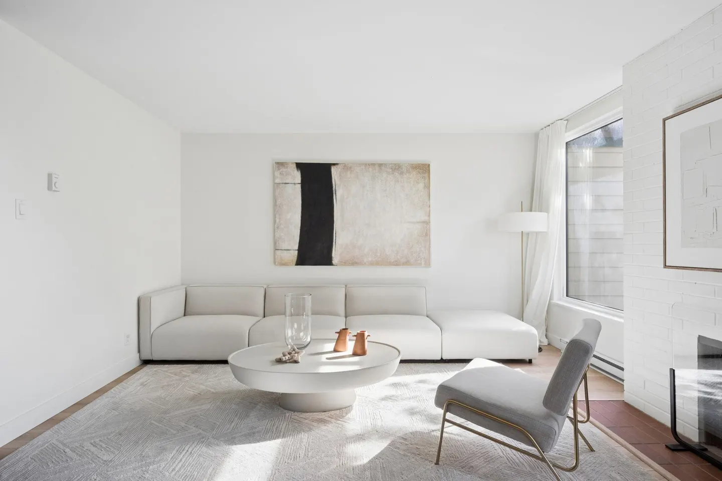 Bright living room with white walls, a sectional sofa, round table, and gray chair. Abstract art hangs above the sofa.