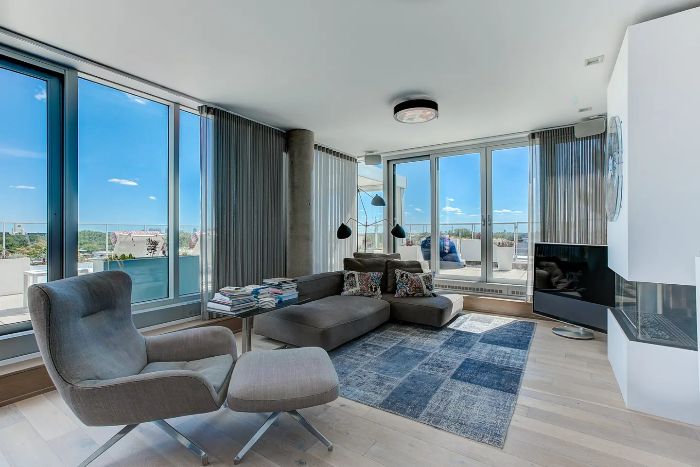 Bright living room with floor-to-ceiling windows, gray sofa, chair, and blue rug. Balcony view of city skyline.