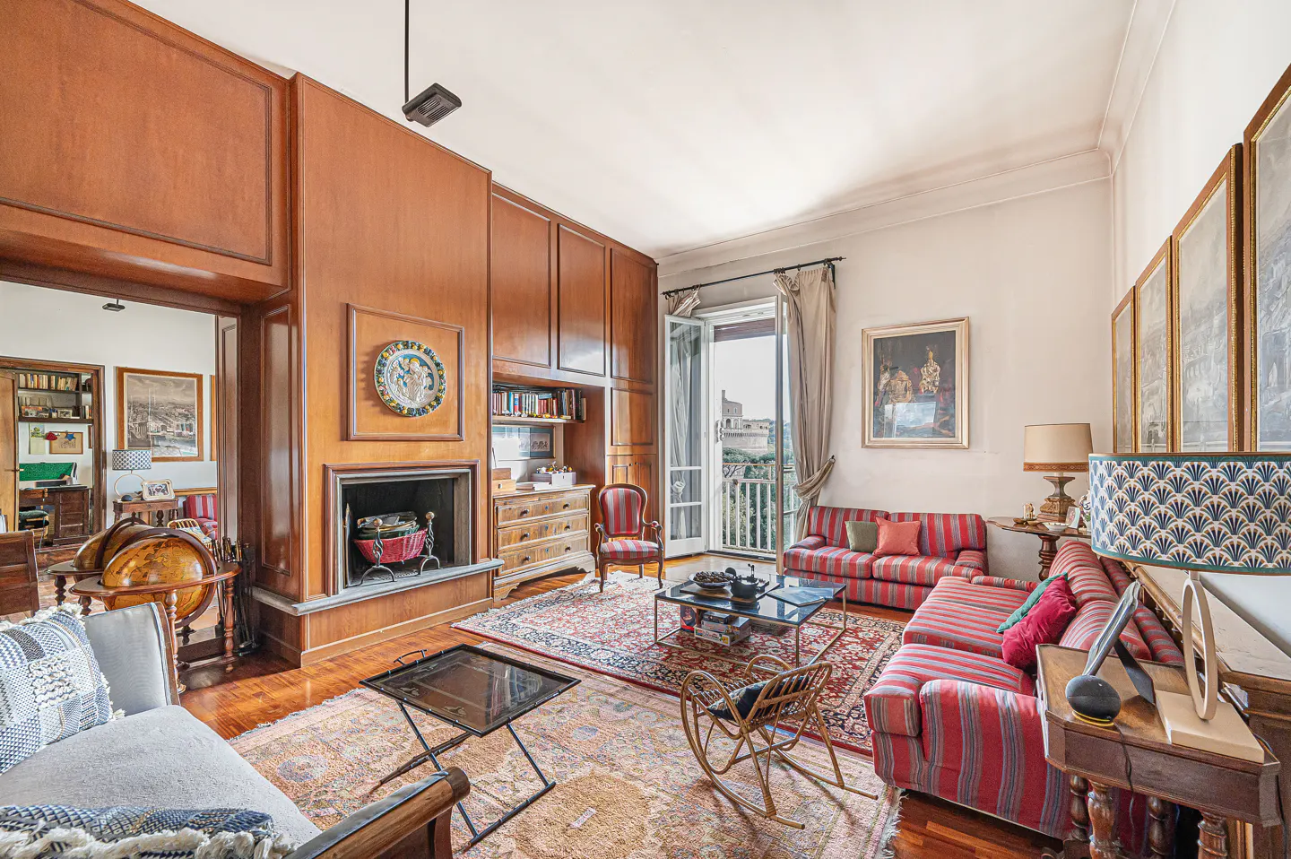 A living room with wood paneling, a fireplace, and a balcony with a view. The room has red and white striped sofas, a patterned rug, and artwork on the walls.