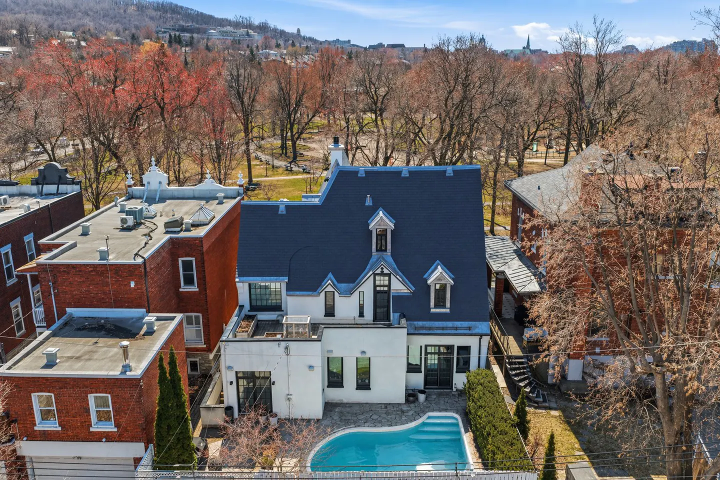 Aerial view of a white house with a blue roof and a pool in the backyard, surrounded by trees and other houses.